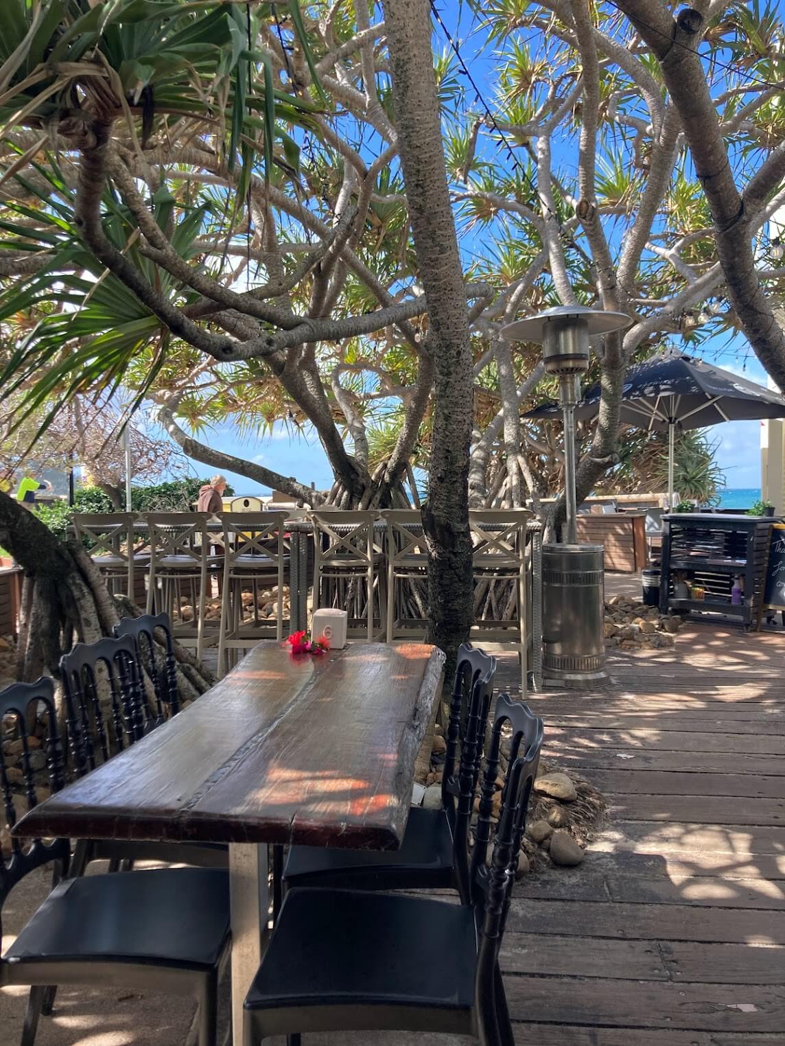 A table and chairs sit outdoors at a cafe surrounded by trees. The ocean is straight ahead
