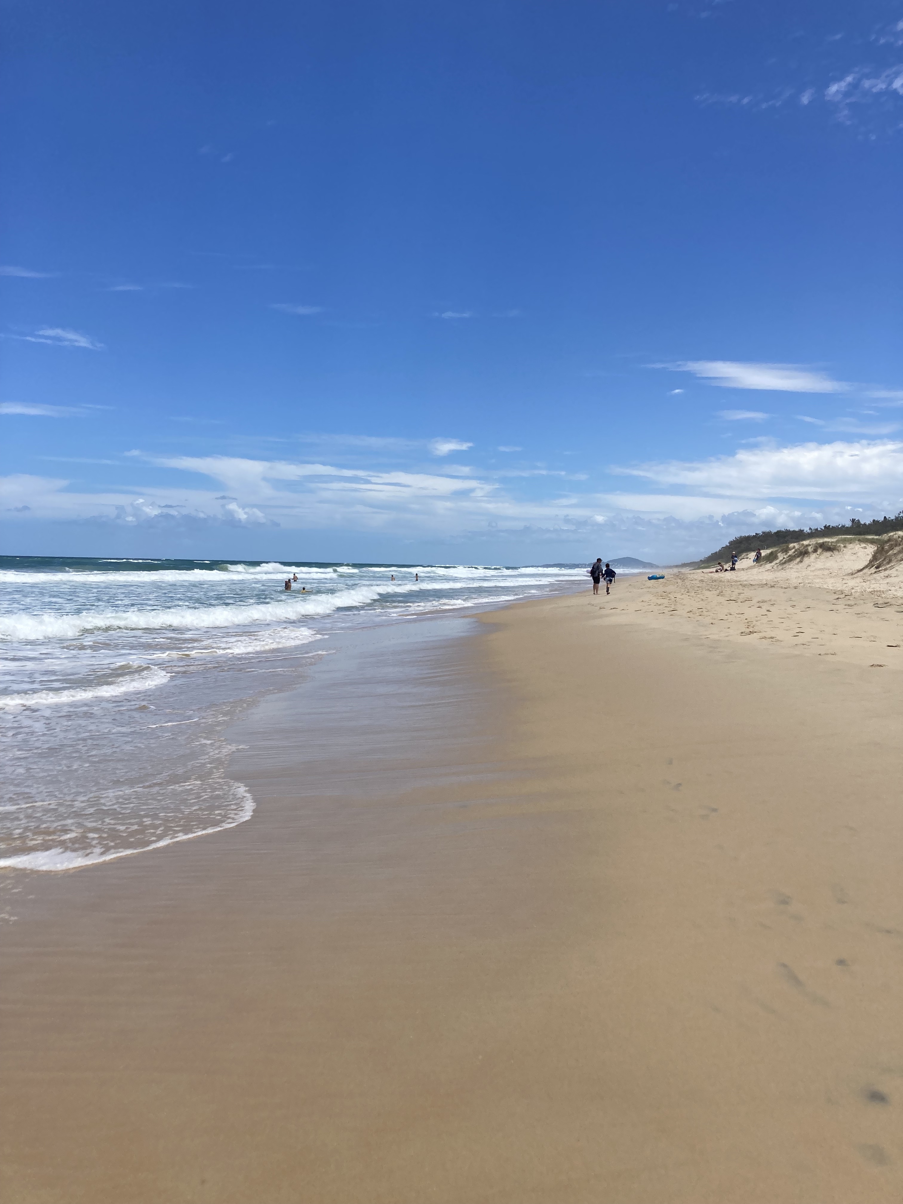 Sunrise beach on a blue sky day with a few white clouds lots of empty sand and a few people in the water