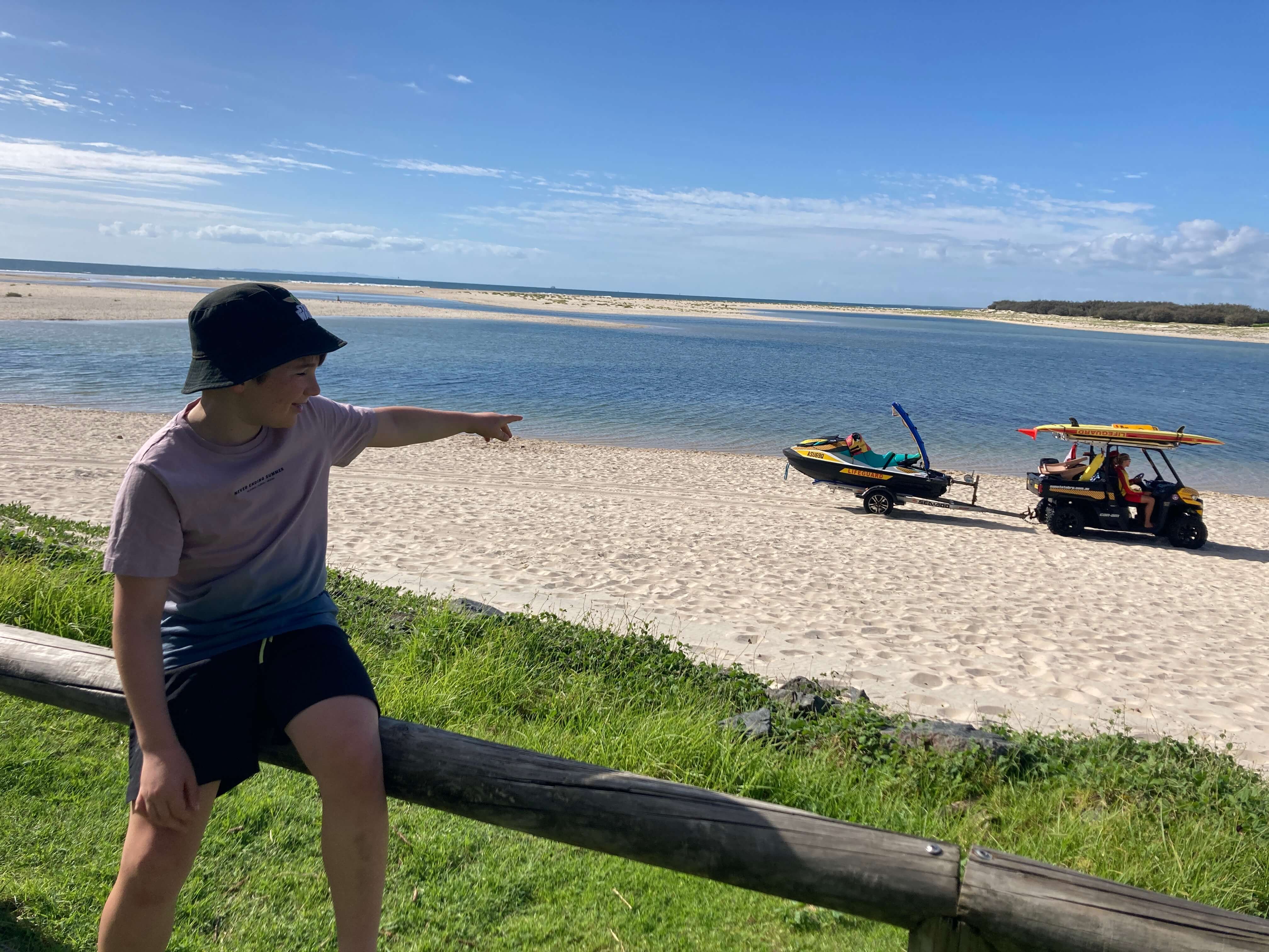 Boy who is sitting on a wooden fence points out lifeguards as they drive past on beach below