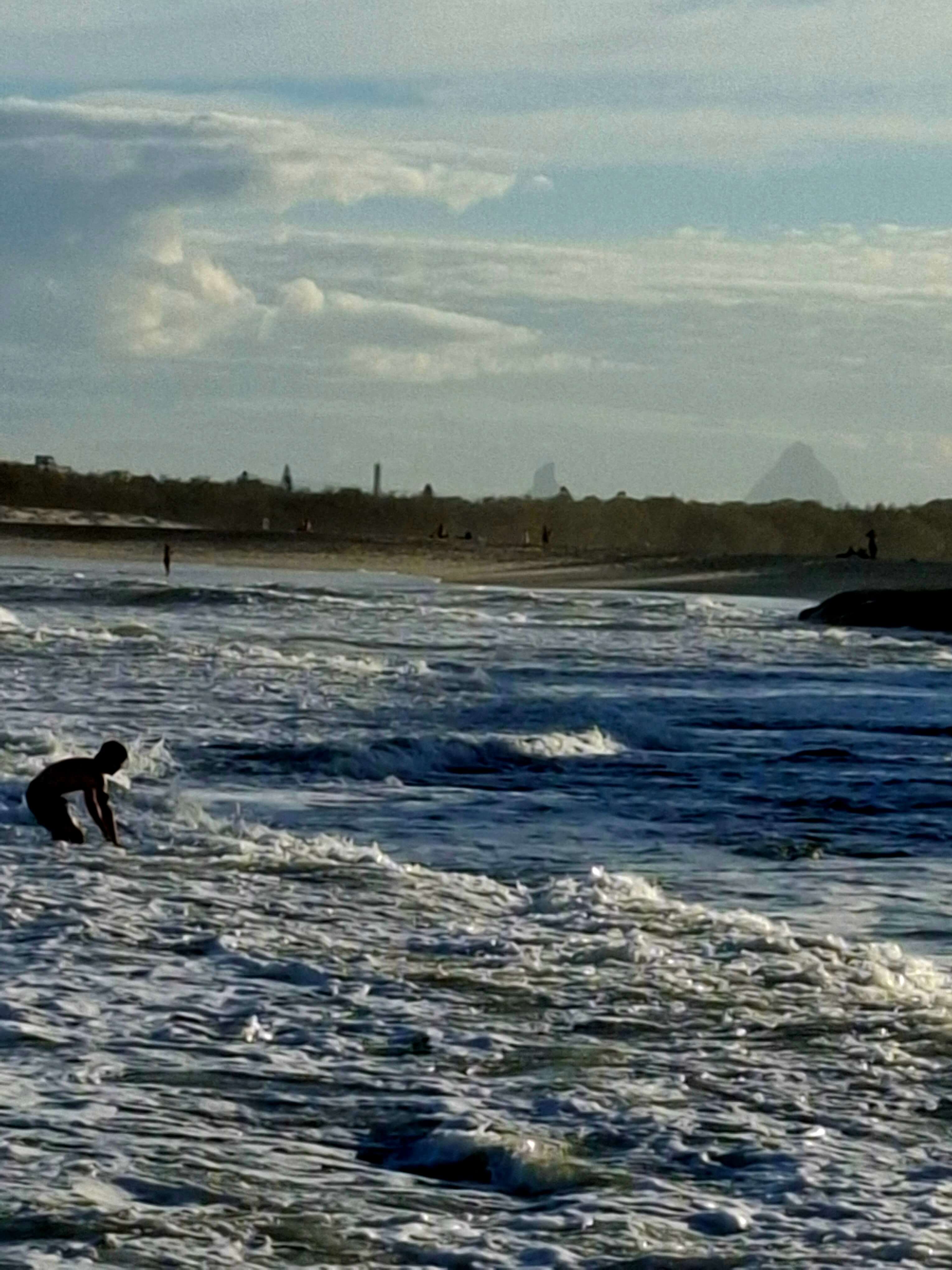 surfer riding wave to shore with glasshouse mountains in background