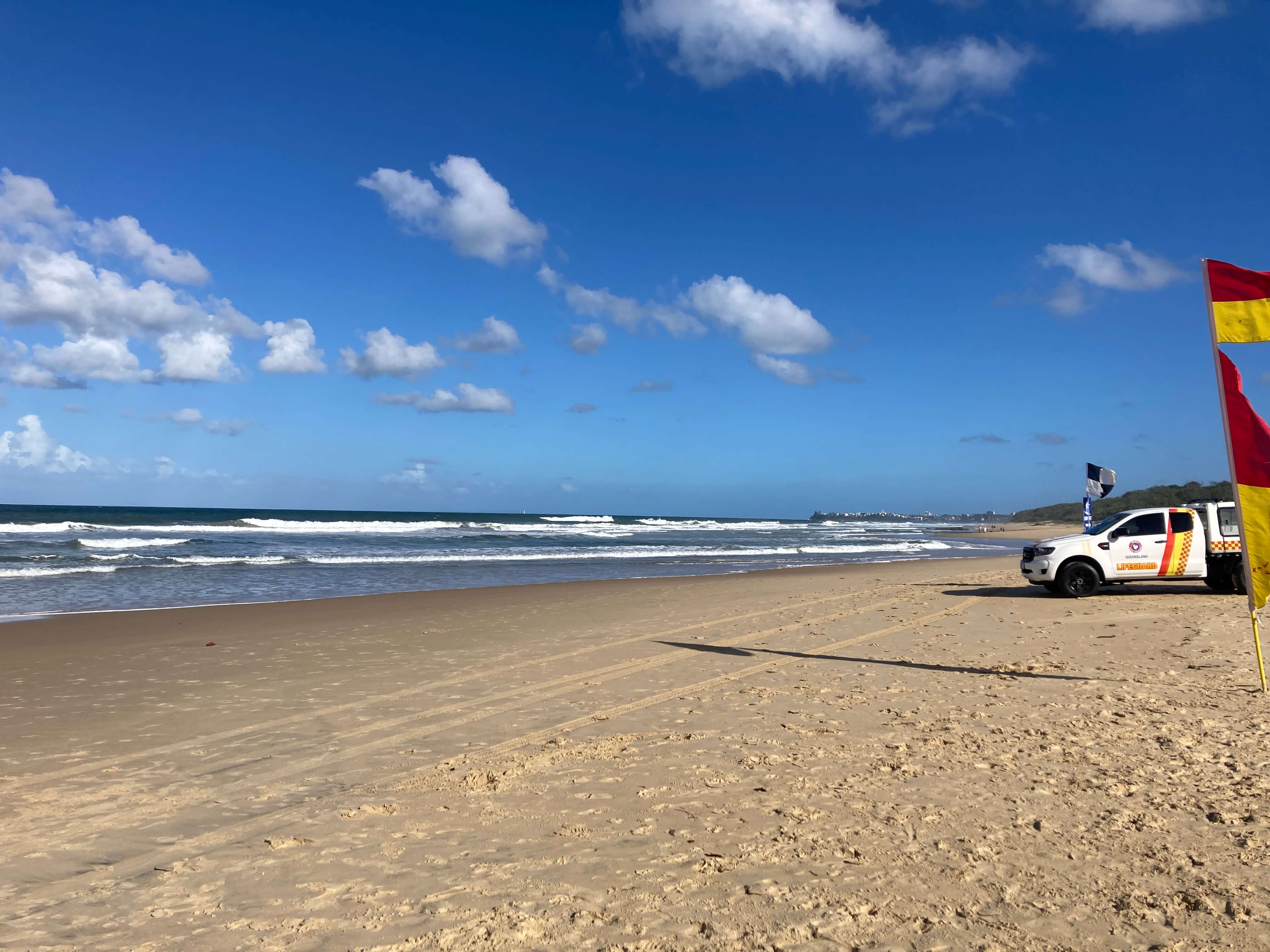 Expanse of beach with a lifeguard vehicle parked on the right hand side of photo. Truck is mainly white. Red and yellow flags are waving in breeze near the vehicle