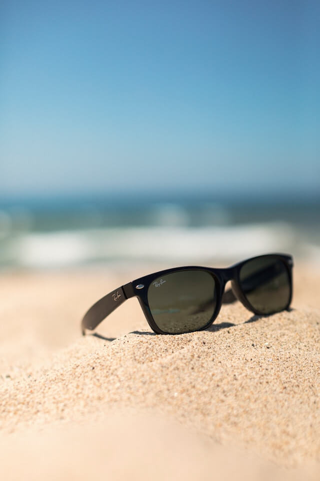 set of sunnies sit on the sand. Water is blurred in the background