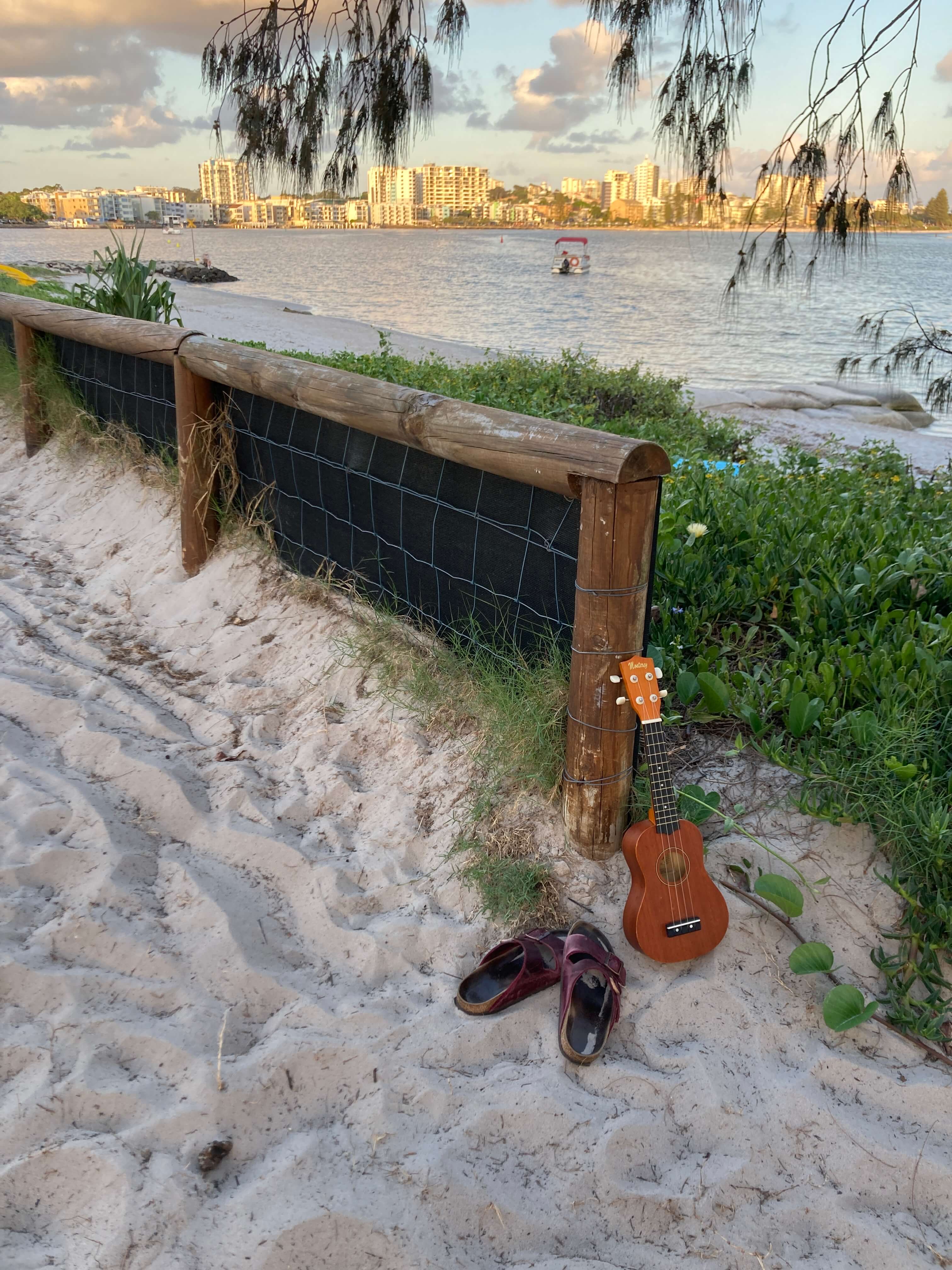 thongs and a ukulele sit on the sand. Town in background with glow of sunset hitting it thongs and a ukulele sit on the sand. Town in background with glow of sunset hitting it